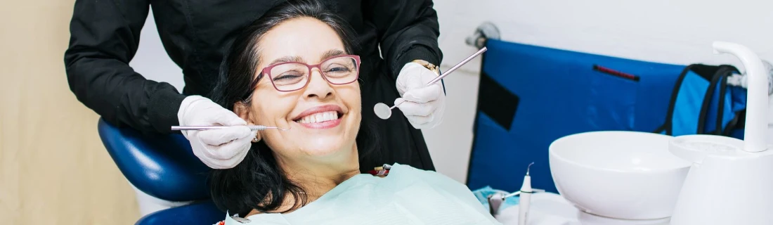 a smiling woman in glasses is leaning back in a dental chair receiving a cleaning